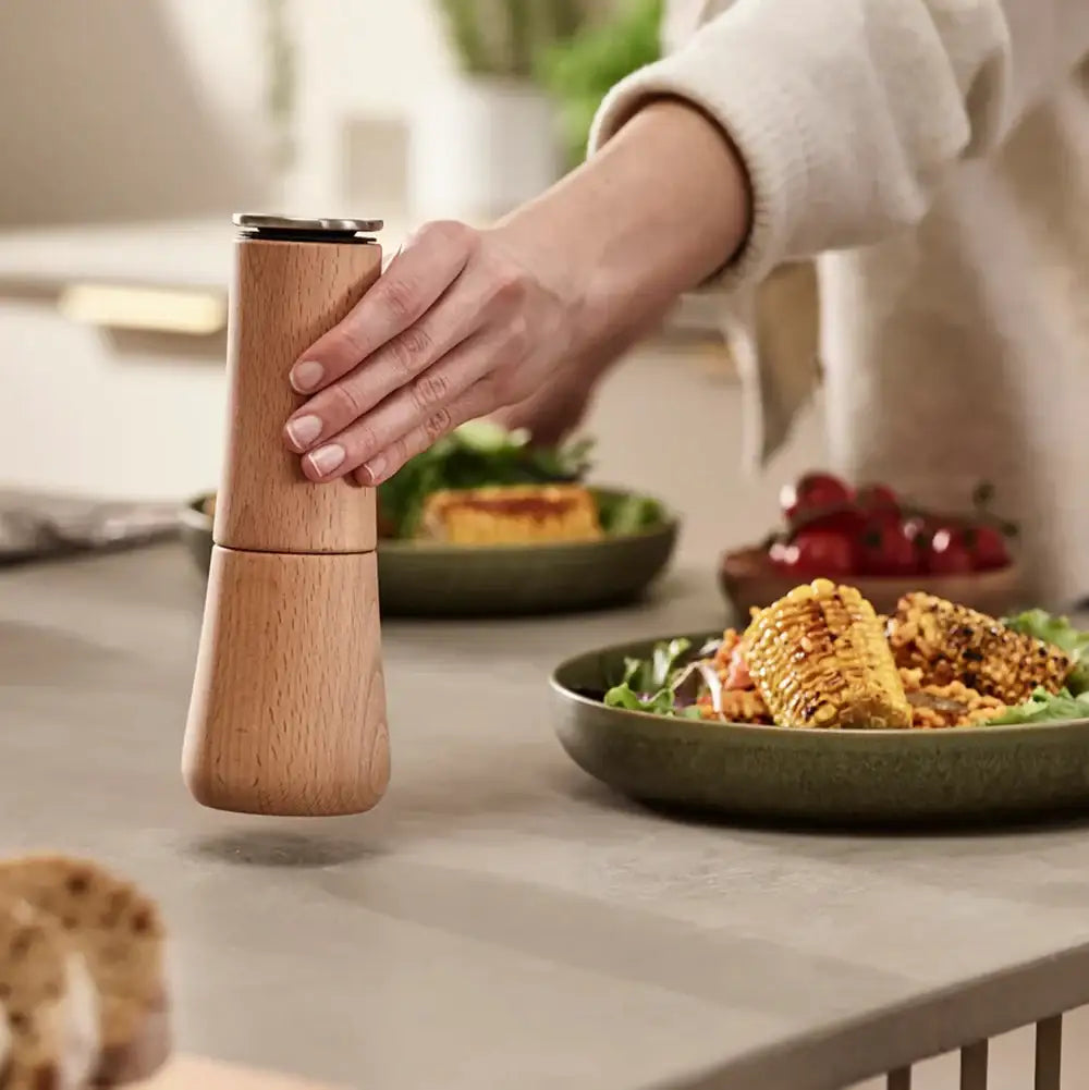 Person using a wooden pepper grinder at a dining table with food.