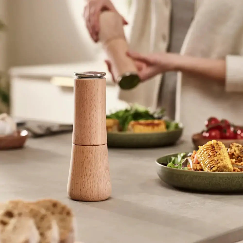 Wooden pepper grinder on a kitchen counter with food in the background