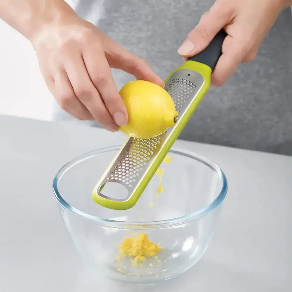 Person using a green citrus zester to zest a lemon over a glass bowl on a white surface.