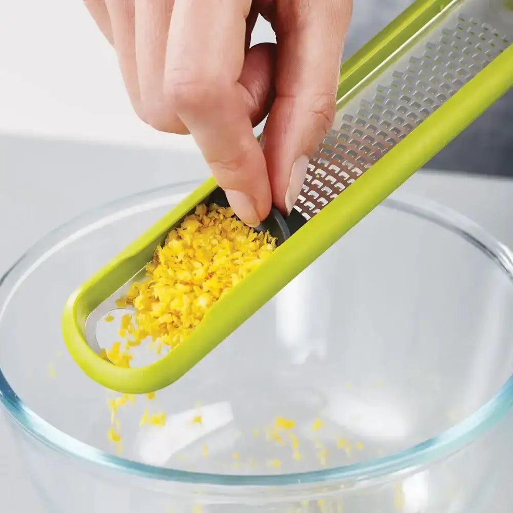 Hand using a green citrus zester to grate yellow zest into a clear glass bowl.