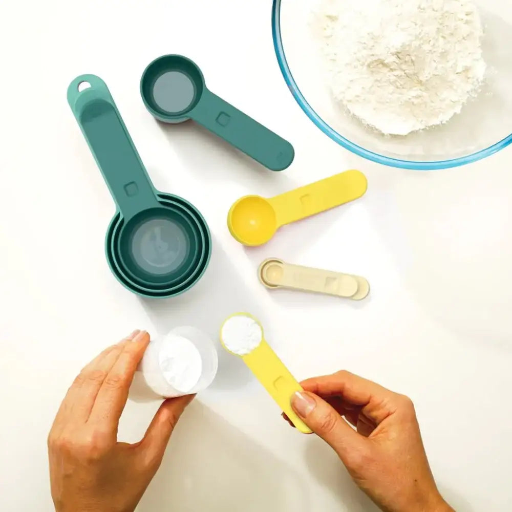 Colorful measuring spoons and cups on a white surface with hands holding a yellow measuring spoon.