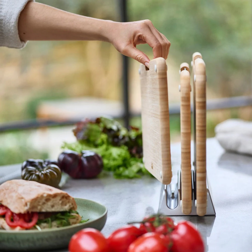 Person reaching for a wooden cutting board on a table with a sandwich and vegetables.