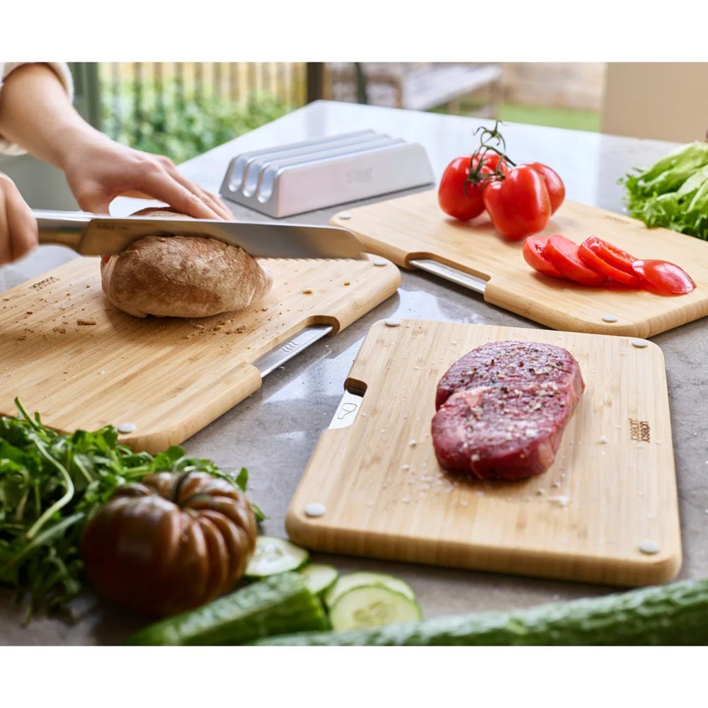 Person slicing bread on a wooden cutting board with vegetables and a steak in the background.