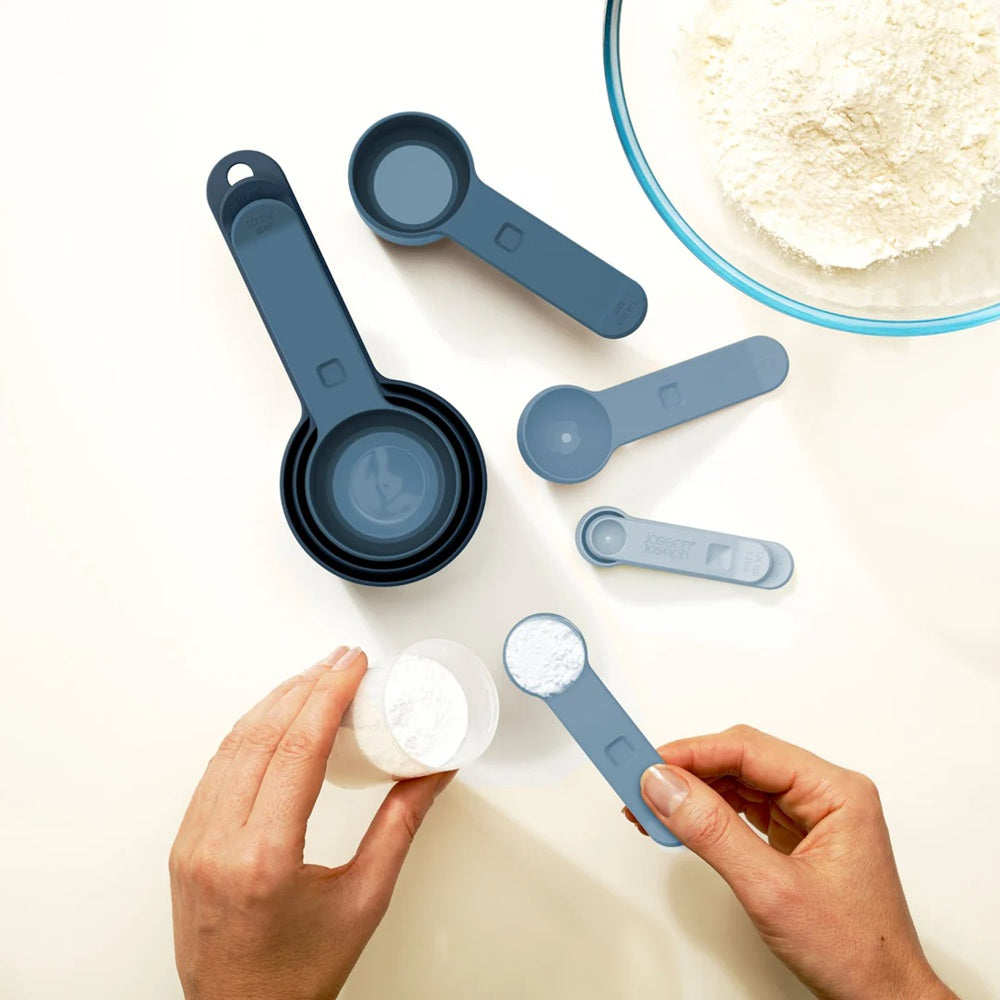 Set of blue measuring spoons and cups on a white surface with hands holding a small container.