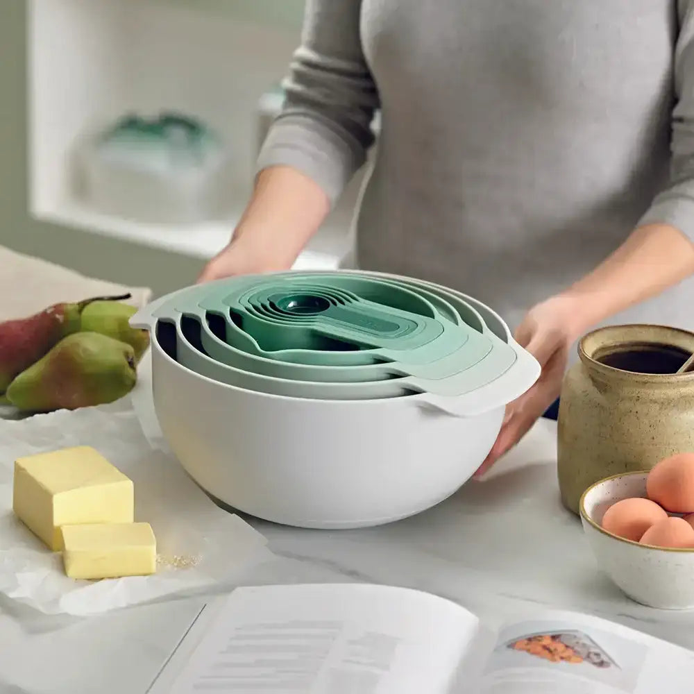 Stack of white and green nesting bowls on a kitchen counter with a person and various items in the background.
