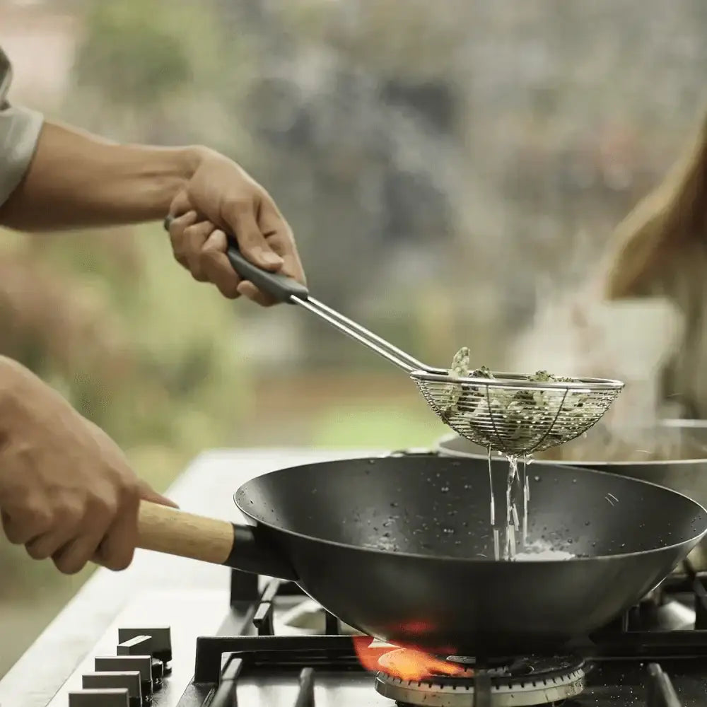 Person cooking outdoors using a wok and straining greens into it.