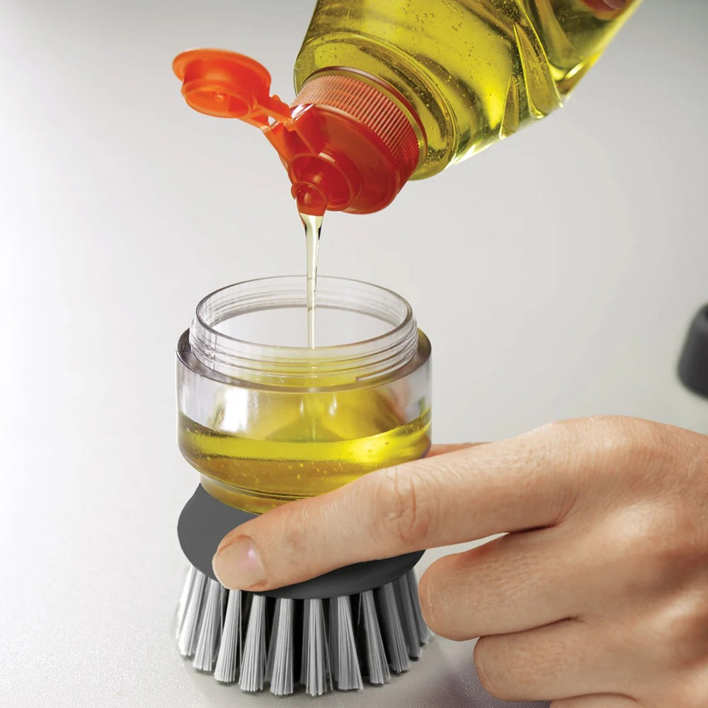Person pouring soap from a bottle into a glass jar with a scrubber at the base.