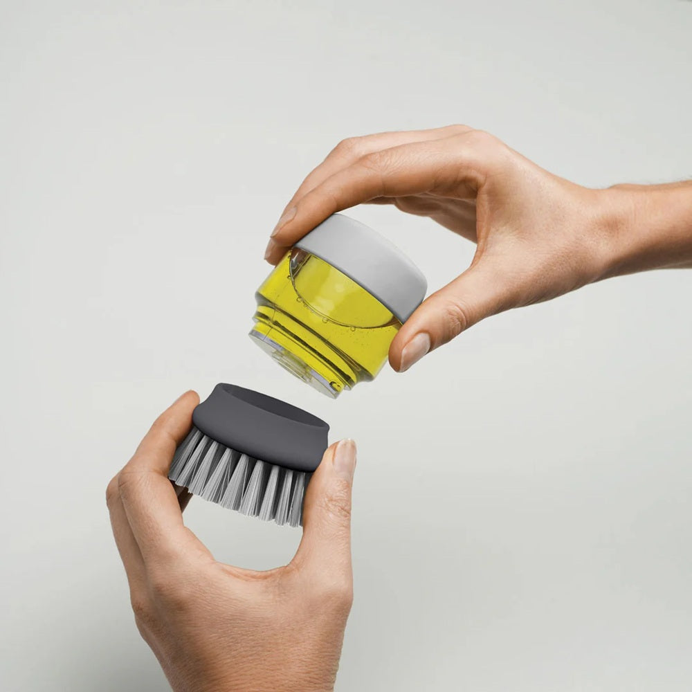 Person holding a gray scrub brush and a clear container with a yellow liquid on a light gray background