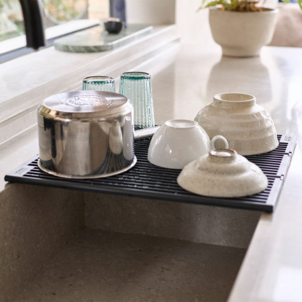 A black silicone dish drainer rack placed over a kitchen sink, holding a stainless steel pot, two blue-green glass tumblers, a white ceramic cup, and two speckled cream bowls.