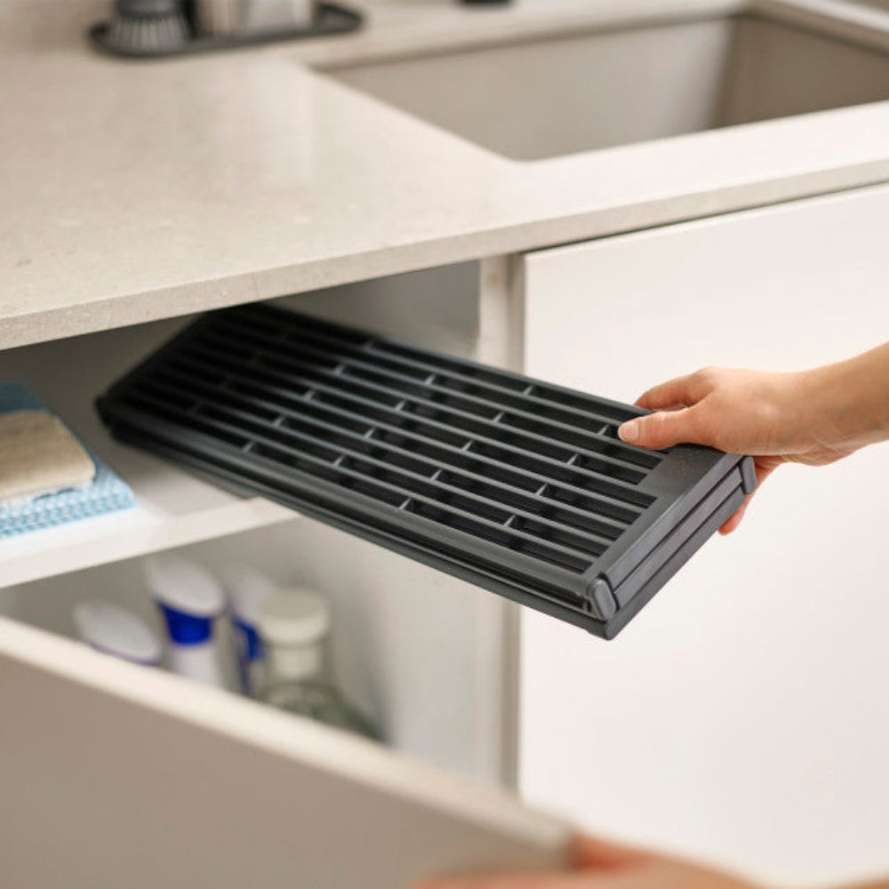 A hand pulls out a black slotted under-sink drawer organizer tray from a white kitchen cabinet.