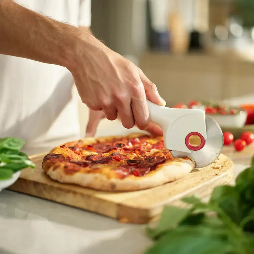 Person using a pizza cutter to slice a pizza on a wooden board with vegetables in the background.