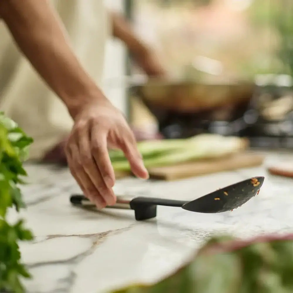 Person holding a black spoon on a kitchen counter with green vegetables.