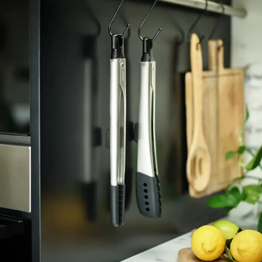 Metallic kitchen tongs hanging on a magnetic strip with lemons and a cutting board in the background.
