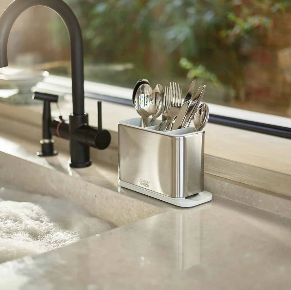 A stainless steel cutlery holder with utensils, set on a beige stone kitchen countertop next to a black faucet and window.