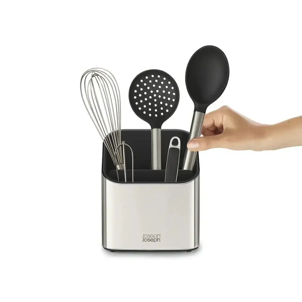 Kitchen utensils in a holder with a hand reaching in to grab a spoon, on a white background.