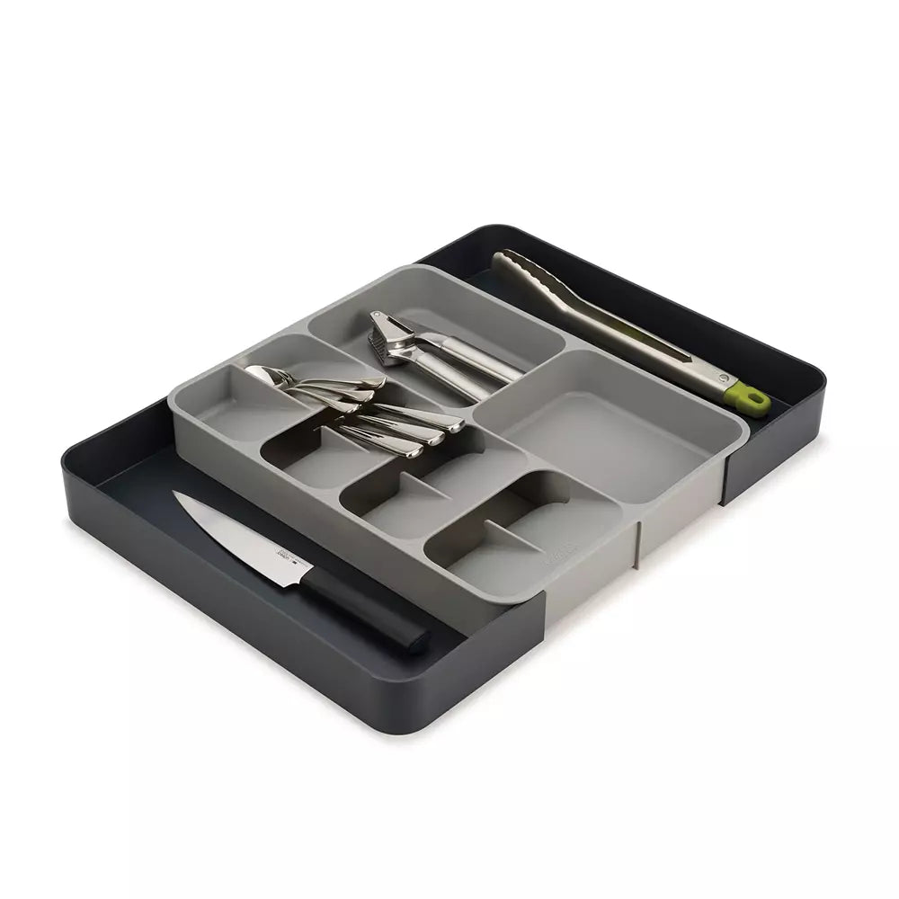 A kitchen utensil organizer tray with a dark charcoal base and a light gray compartment insert, holding several stainless steel tools including tongs, a small knife, and a spoon, on a white background.