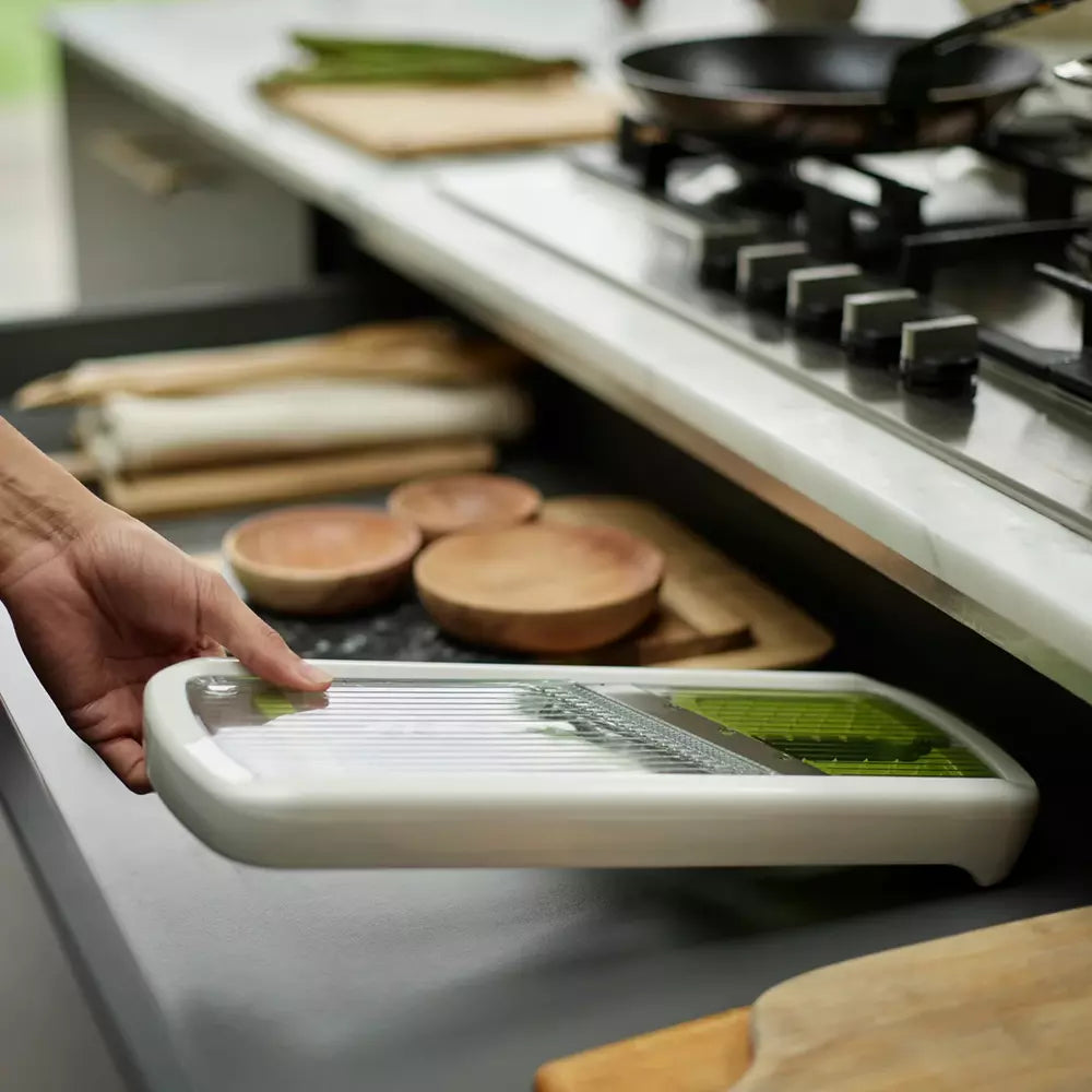 A hand is sliding a white mandoline slicer with green vegetables on a dark kitchen countertop, with a stove and kitchen items in the background.