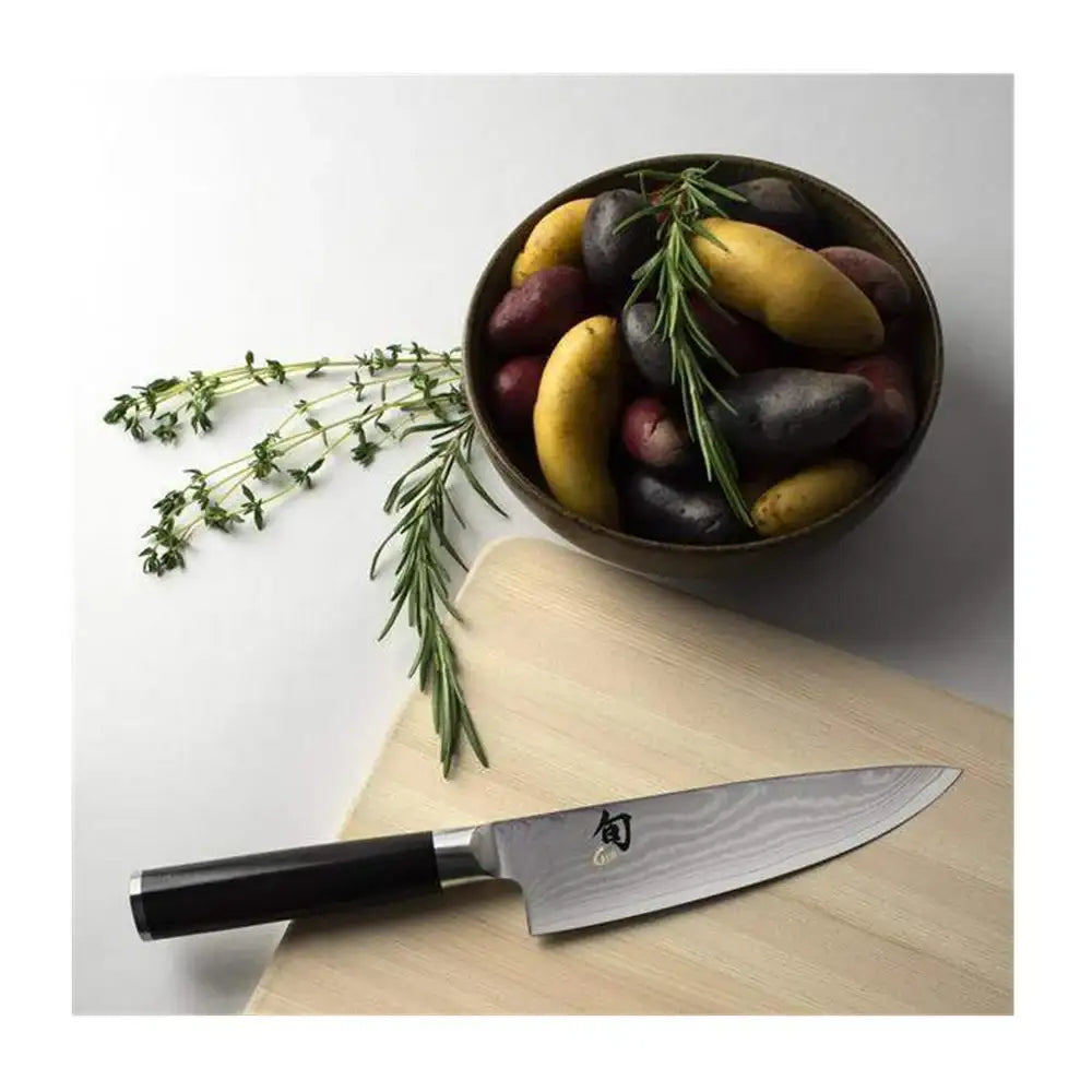 Chef's knife on a wooden cutting board with a bowl of vegetables and herbs in the background.