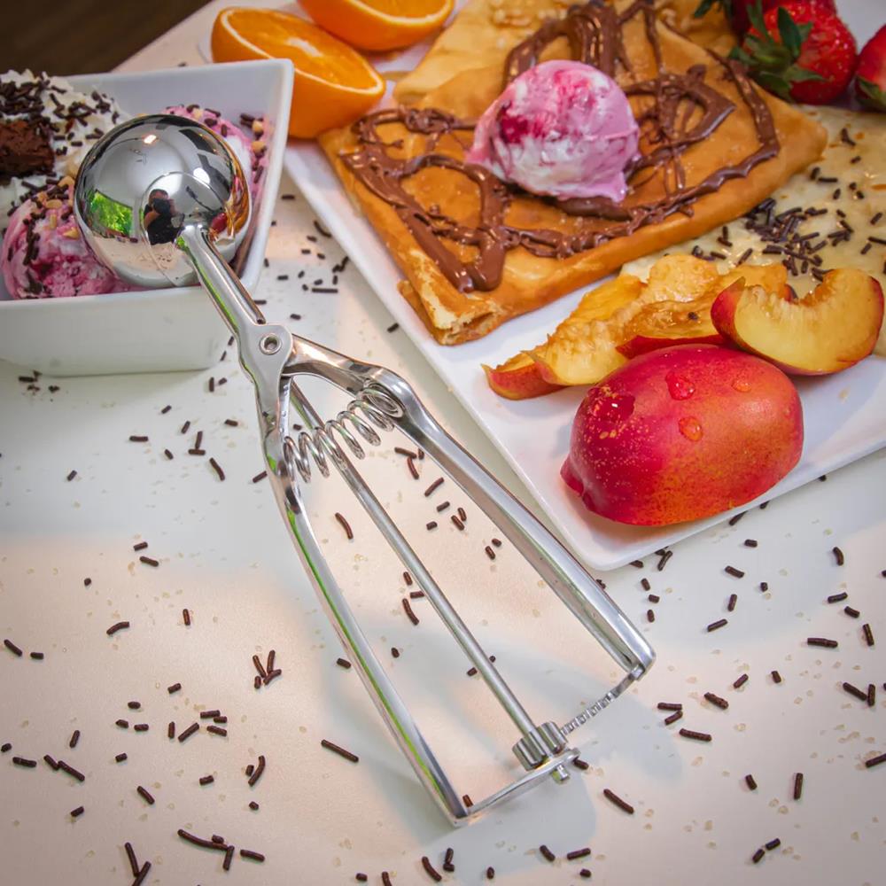 Silver ice cream scoop on a table with dessert items including waffles, fruit, and ice cream.