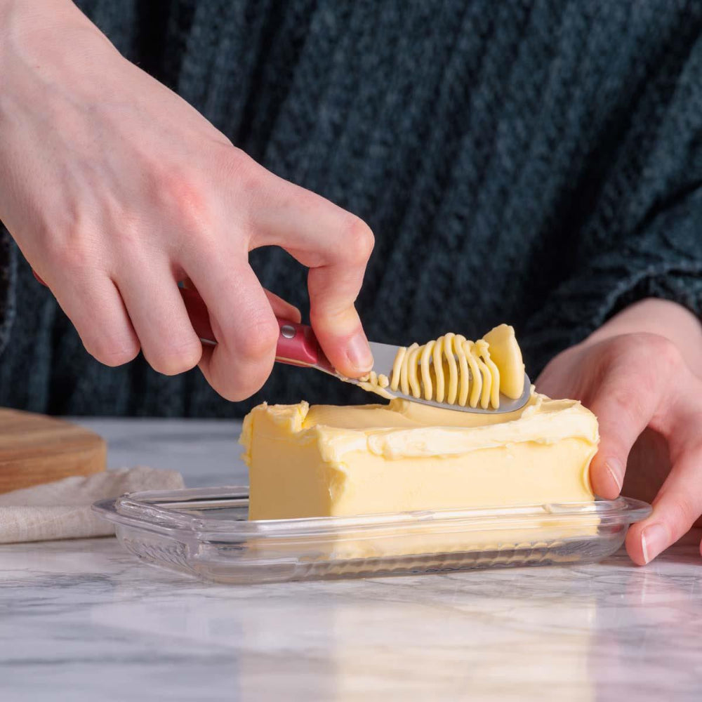A close-up of a person's hands using a butter knife to spread softened butter on a stick in a clear plastic tub, with a dark textured sweater visible in the background.