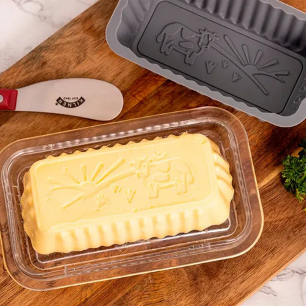 A rectangular yellow butter mold in a clear plastic container, shown with a gray metal mold and a spatula on a wooden cutting board.