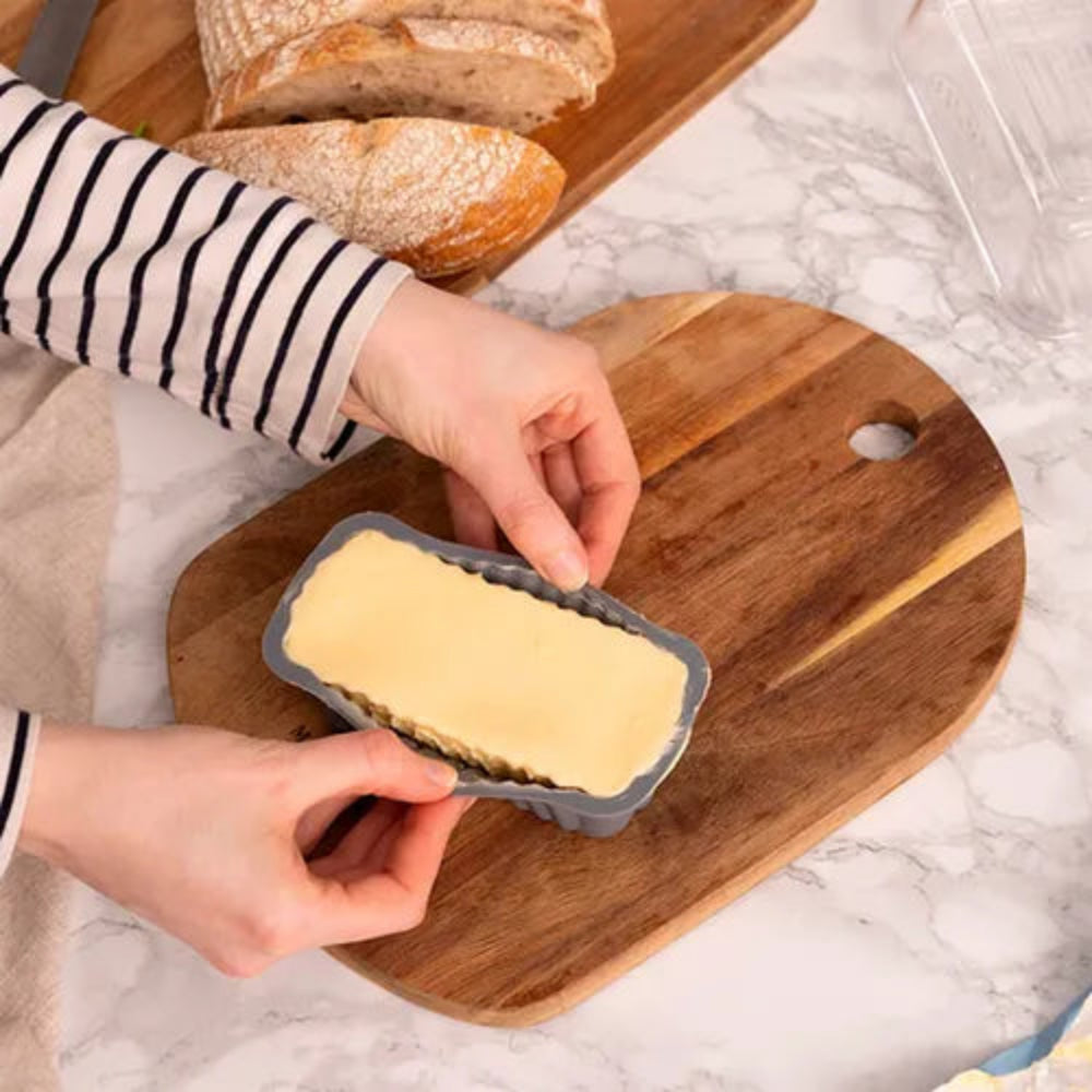 A person wearing a black-and-white striped long-sleeve shirt holds a gray rectangular loaf pan filled with pale yellow dough or batter, placed on a wooden cutting board over a white marble surface. Sliced bread is visible in the background on another wooden board.