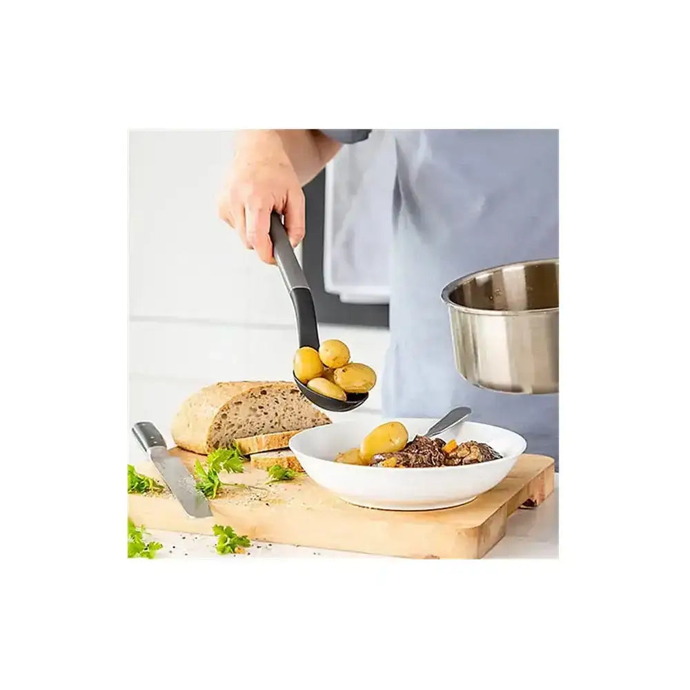 Person holding a ladle over a bowl of food on a wooden cutting board with bread and olives.