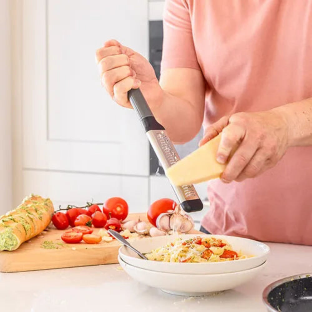 A person in a pink shirt grates cheese into a bowl of pasta in a bright kitchen. The cheese grater has a black handle and a silver metal blade with the brand name 'Kitchenaid' visible.