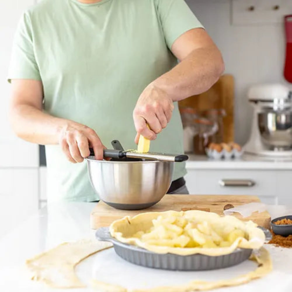 A person in a light green t-shirt grates cheese into a stainless steel mixing bowl on a wooden cutting board, with a pie crust and apple filling in the foreground and a kitchen stand mixer in the background.