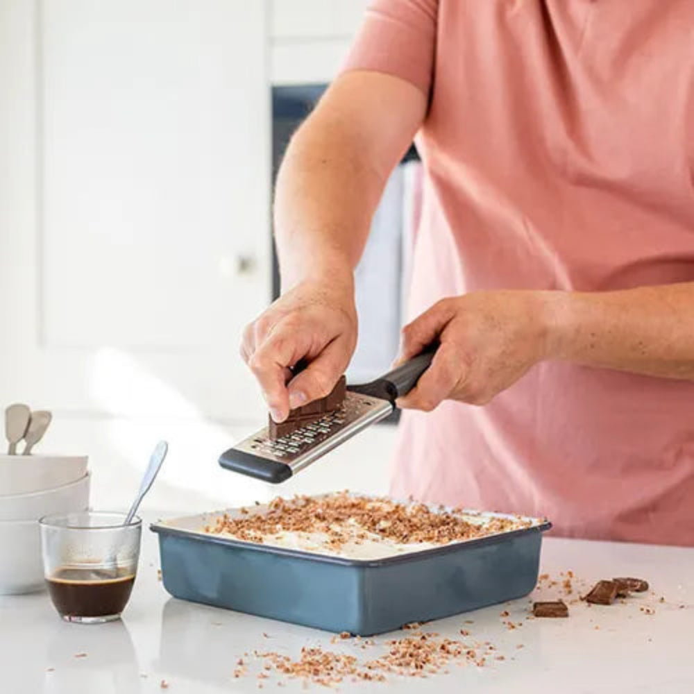A person in a pink shirt grates chocolate onto a dessert in a square blue baking pan on a white countertop in a kitchen.