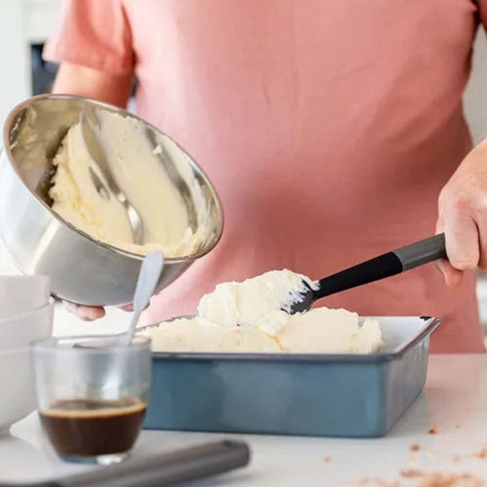 A person in a pink shirt transfers creamy white frosting from a stainless steel mixing bowl into a blue rectangular baking pan using a black spatula, with a small glass of espresso in the foreground.