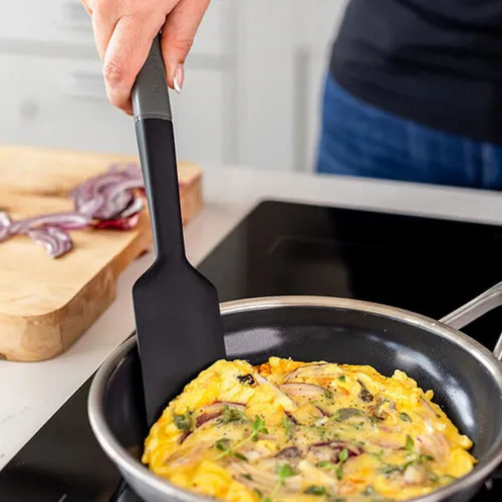 A black silicone spatula with a gray handle being used to flip an omelet in a black frying pan on an induction cooktop, with a person's hand and torso visible in a kitchen setting.