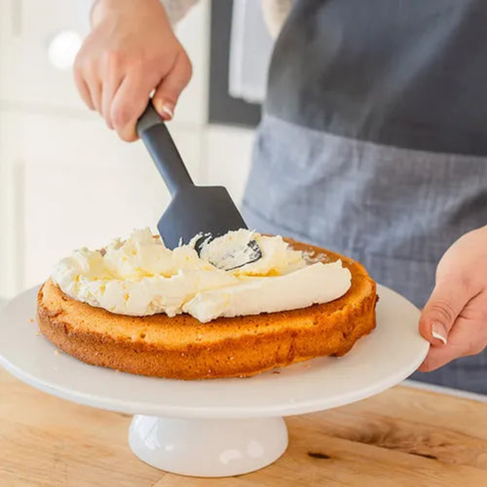 A person in a gray apron uses a black spatula to spread white frosting onto a round cake layer on a white cake stand, set on a wooden table.