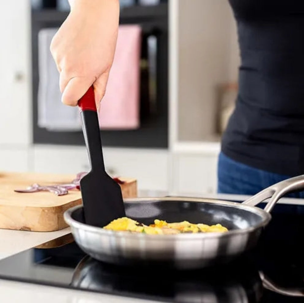 A person in a kitchen uses a black silicone spatula with a red handle to flip food in a stainless steel frying pan on a black stovetop.