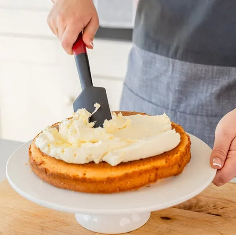 A person in a gray apron uses a spatula with a red handle to spread white frosting onto a round cake layer on a white cake stand, set on a wooden surface.