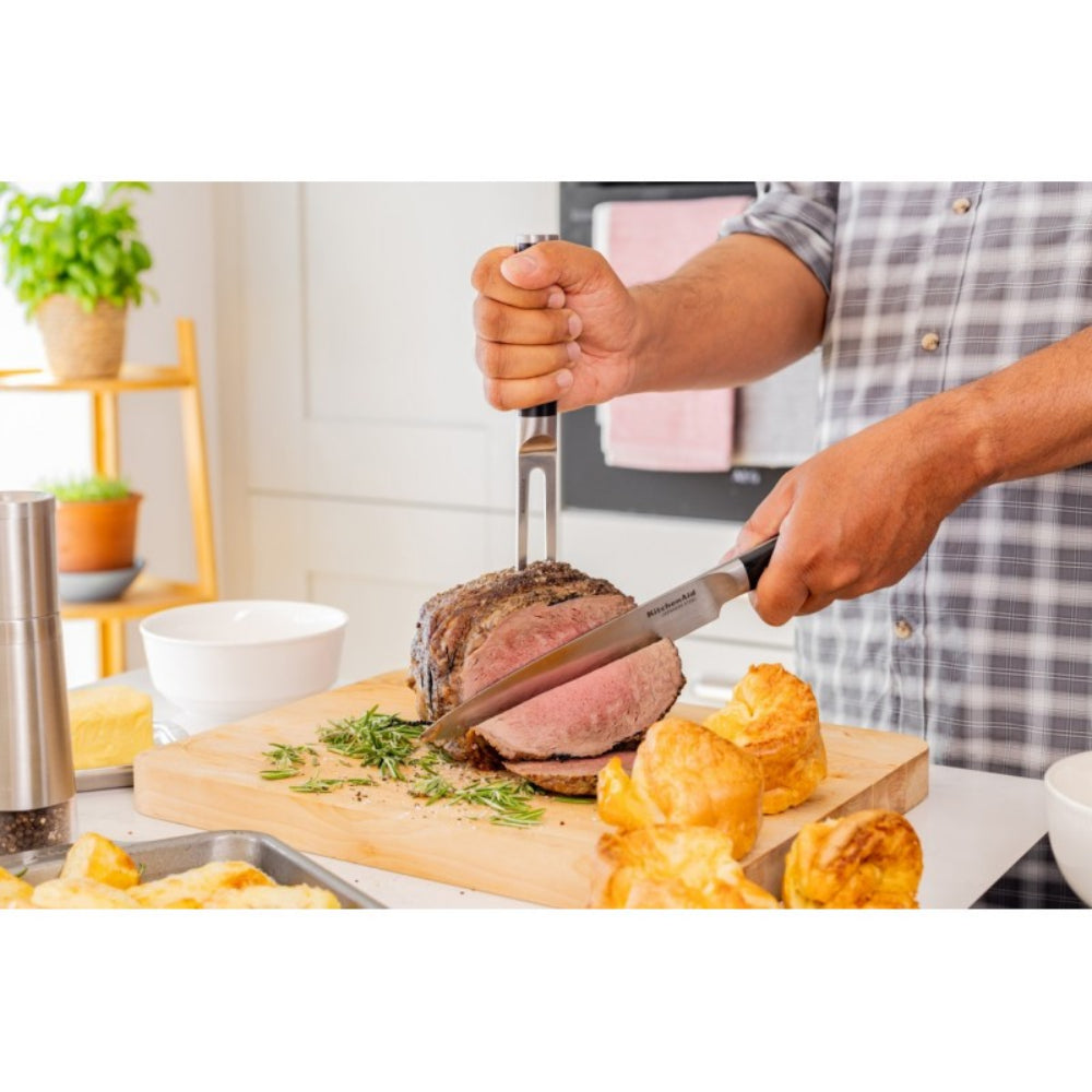 A person slicing a cooked beef roast on a wooden cutting board with a carving knife and fork, with roasted potatoes and kitchen items in a bright kitchen setting.