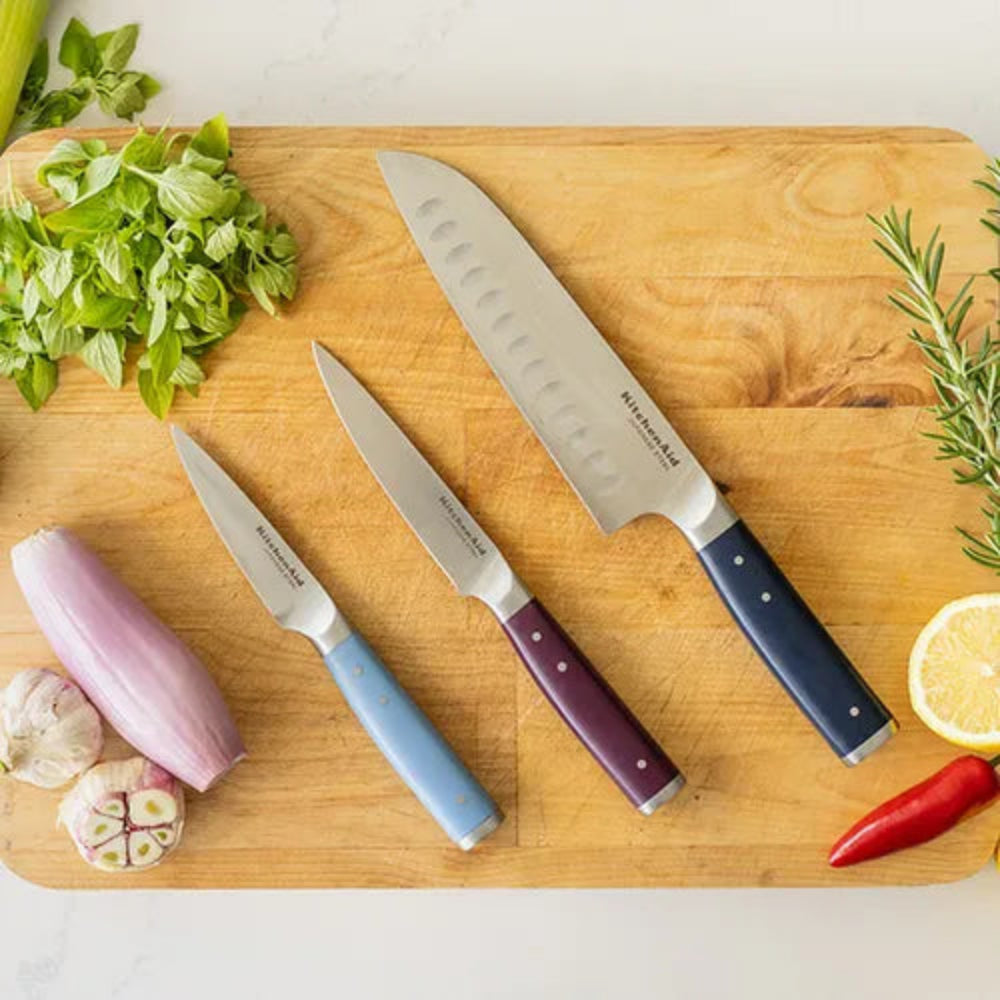 Three Mikasa kitchen knives with colorful handles in blue, purple, and navy are arranged on a wooden cutting board alongside fresh ingredients including basil, shallot, garlic, lemon, rosemary, and chili.