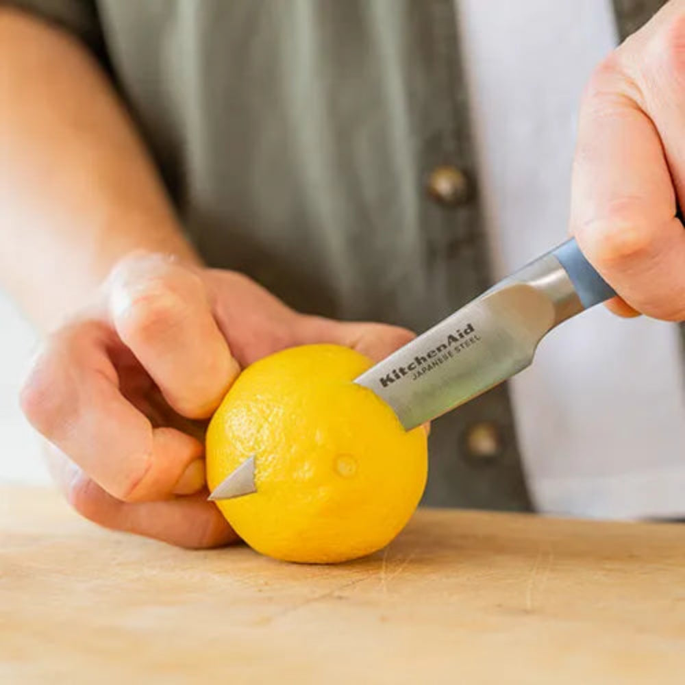 A close-up of a person using a KitchenAid Sharpie utility knife to cut a lemon on a wooden cutting board. The person is wearing a gray button-up shirt.