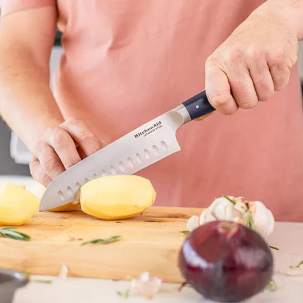 A person in a pink apron slicing a peeled yellow potato with a KitchenAid Santoku Series kitchen knife on a wooden cutting board, with garlic, rosemary, and a red onion nearby.