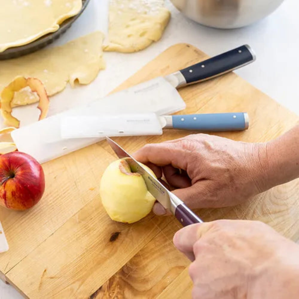 A close-up lifestyle shot of a person's hands using a kitchen knife to cut a yellow apple on a wooden cutting board. A white plastic peeler and another knife with a blue handle are also visible on the board, alongside a red apple and pastry dough in the background.