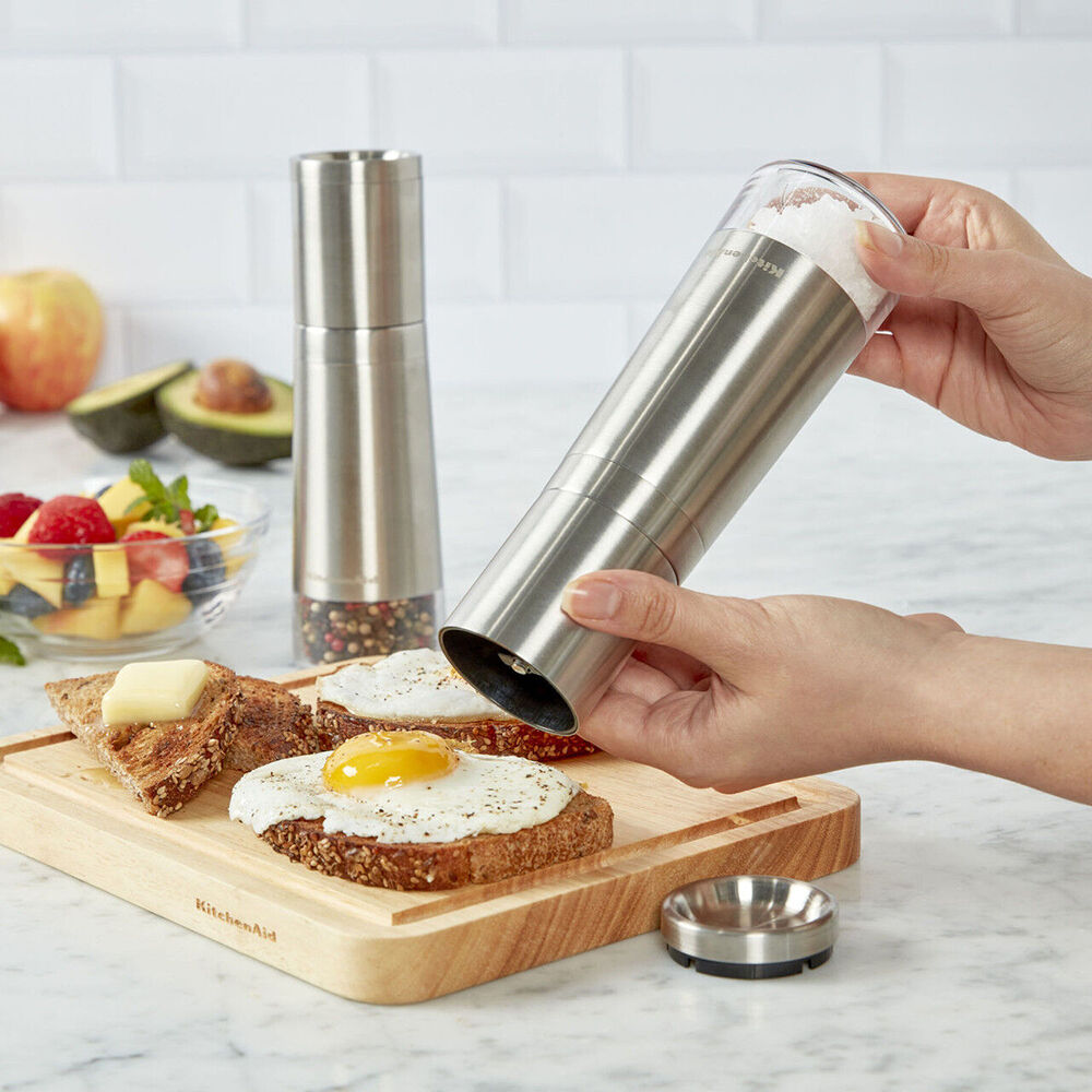 Person using a pepper grinder on toast with an egg, surrounded by fruits and a pepper mill.