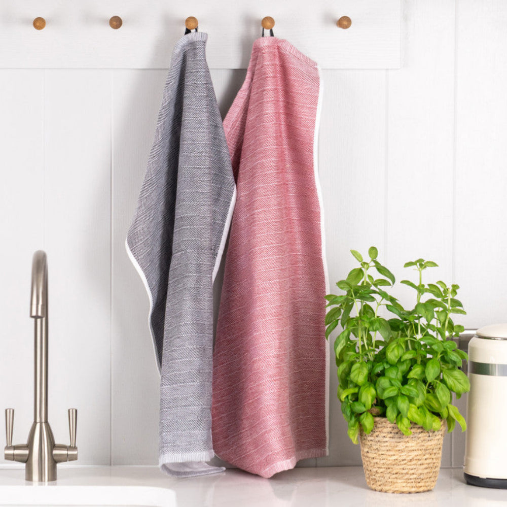 Two textured kitchen towels, one gray and one dusty pink, hanging from wooden wall hooks above a sink. A potted green plant and a cream kettle sit on the counter to the right against a white paneled wall.