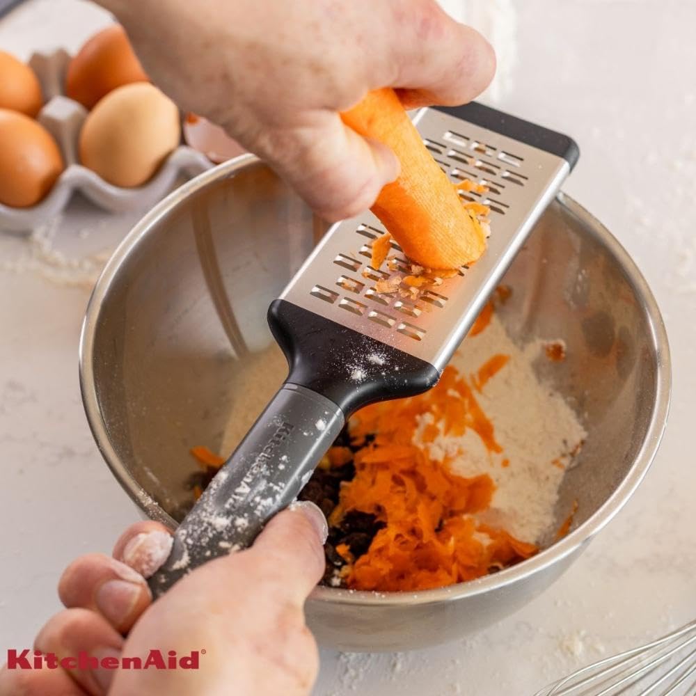 Person using a KitchenAid grater to shred carrots into a bowl.