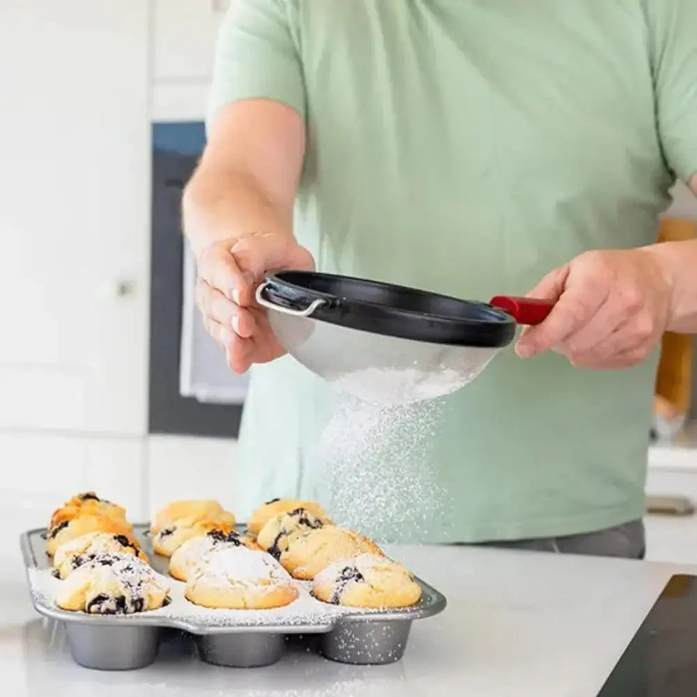 Person dusting powdered sugar over muffins using a small sieve in a kitchen.