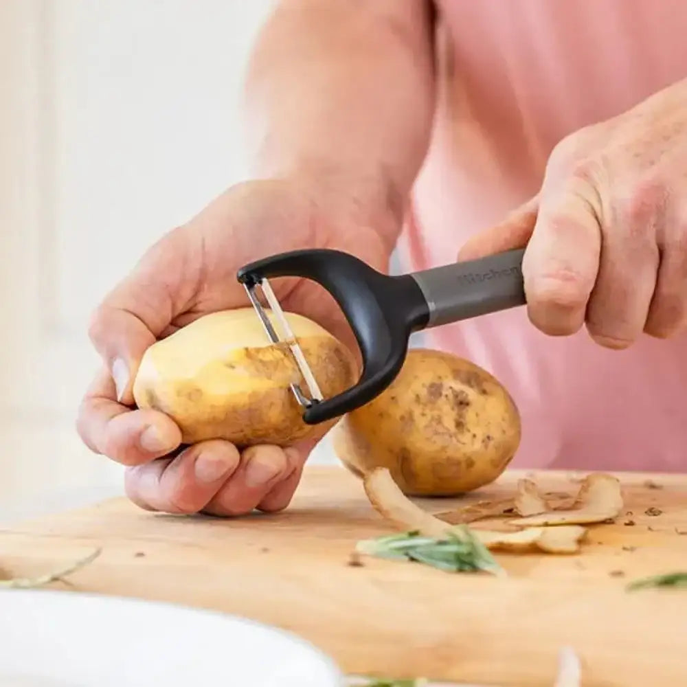 Person peeling a potato with a black and grey peeler on a wooden cutting board.