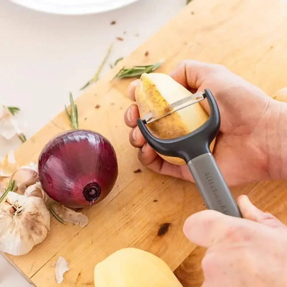 Person peeling a potato with a Y-shaped peeler on a wooden cutting board.