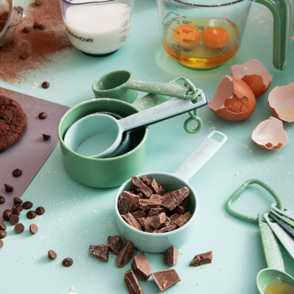 A styled flat-lay of mint green measuring cups and measuring spoons on a light blue-green surface, surrounded by baking ingredients including chocolate chunks, milk, eggs, cracked eggshells, cocoa powder, and nuts.