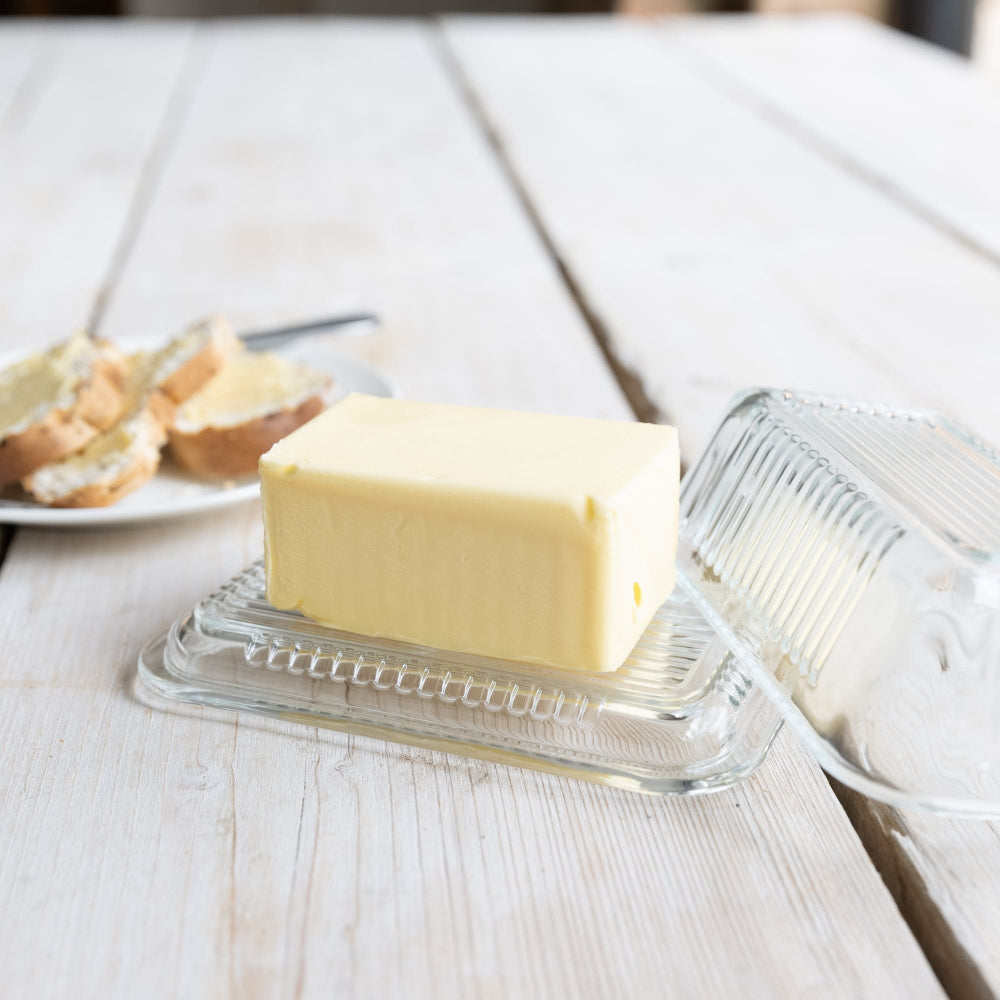 A rectangular block of pale yellow butter sits on a clear ribbed plastic butter dish, with the lid leaning beside it on a light wood tabletop. A plate with bread and a knife is blurred in the background.