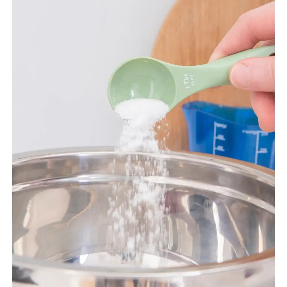 Person pouring salt from a green measuring spoon into a metal bowl.