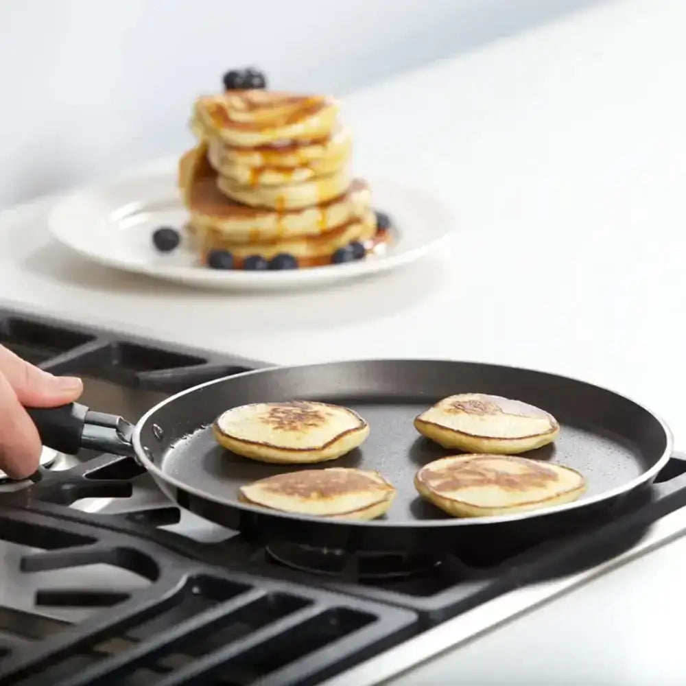 Person cooking pancakes on a griddle with a stack of pancakes and blueberries in the background.
